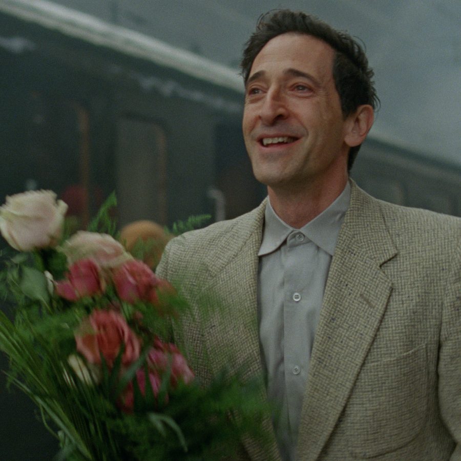  In a scene from The Brutalist, László Tóth (played Adrien Brody) stands on a train station platform, smiling and holding a bunch of flowers.